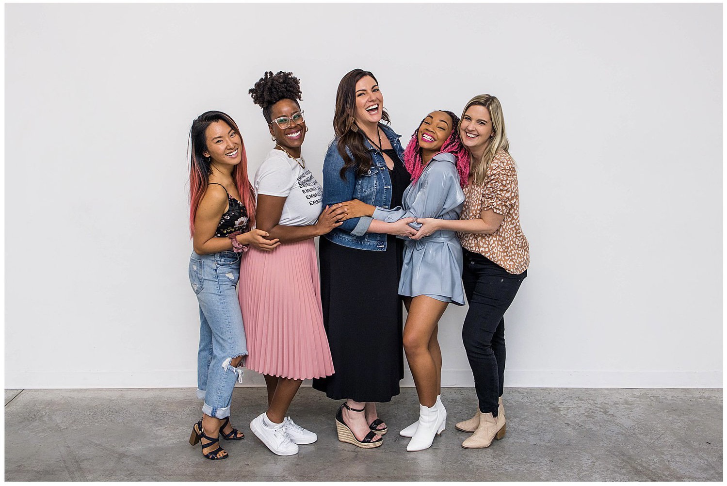 Group of five diverse women, Former DCA Students standing and smiling together with Amy Porterfield in front of a white wall, radiating joy and connection. This image represents the supportive community and success stories highlighted in the Digital Course Academy Review 2025.