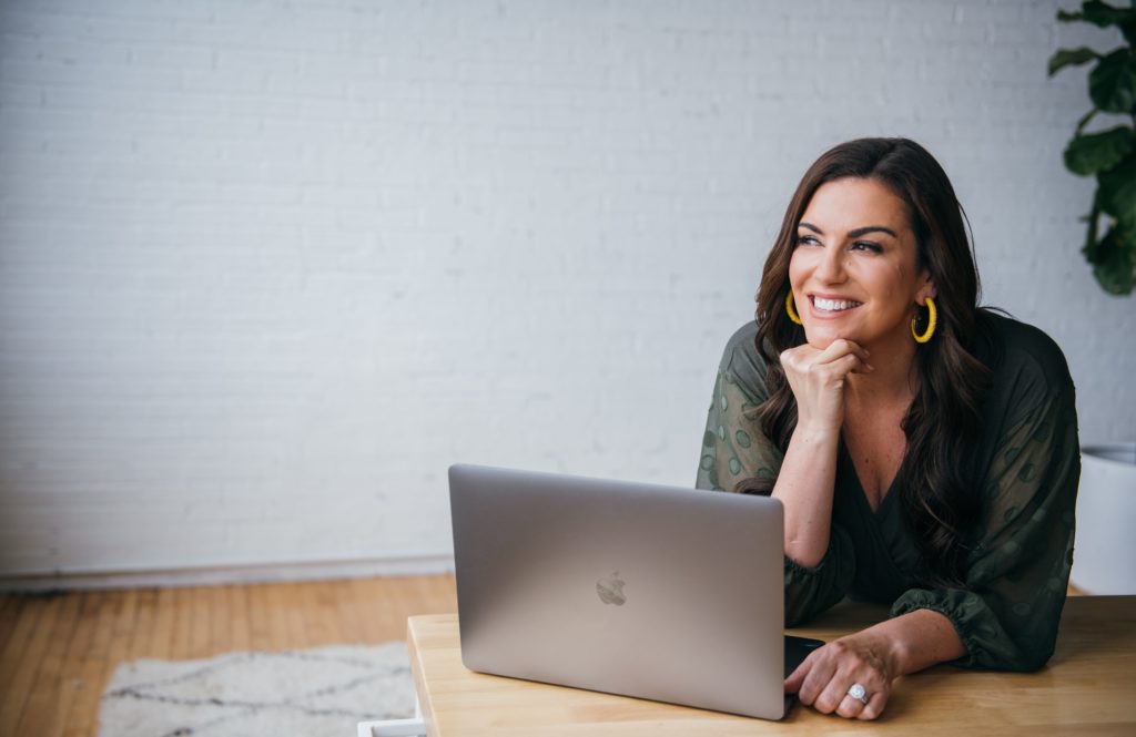 Amy Porterfield smiling while seated at a desk with a laptop, symbolizing the approachable and empowering vibe of her Digital Course Academy (DCA) 2025 program.