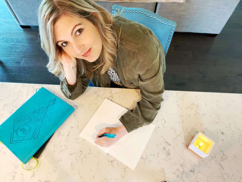 Fempreneur Jamie Trull writing in a notebook at a marble desk, with a candle and bright blue planner nearby. The scene captures a quiet moment of focus, reflection, and planning.