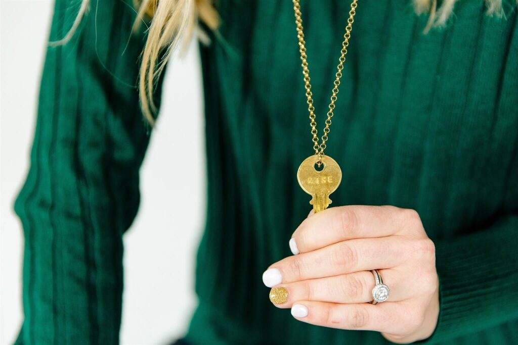 Close-up of fempreneur Jamie Trull wearing a green sweater, holding a gold key necklace engraved with the word "RISE"—symbolizing empowerment and growth for female entrepreneurs.