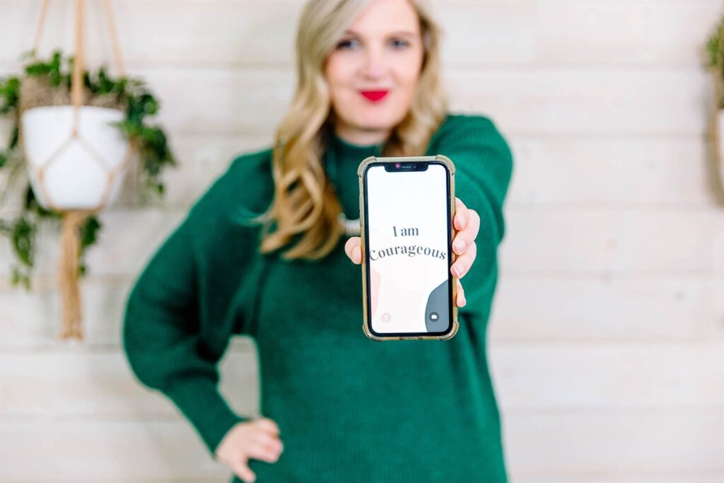 Woman in a green sweater confidently holding a phone that displays the affirmation “I am courageous,” symbolizing empowerment in online education. The image reflects the mindset needed to define learning objectives and succeed in creating an online course.
